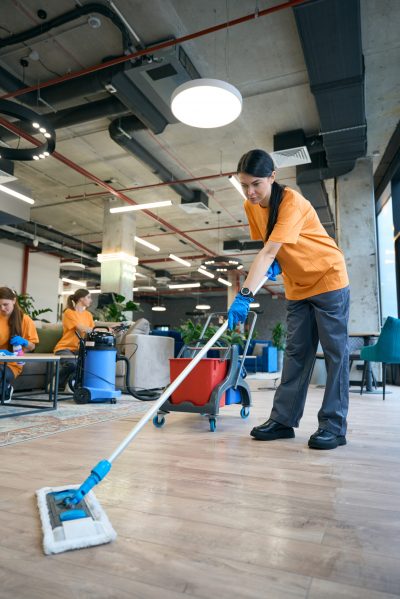 A woman dressed in an orange shirt cleans the floor with a mop, exemplifying a cleaning service in Brisbane.